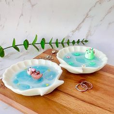 two white dishes filled with blue liquid on top of a wooden cutting board next to a green plant