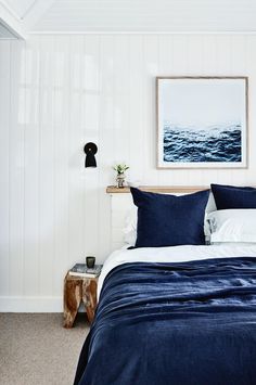 a bed with blue comforter and pillows in a white room next to a painting on the wall