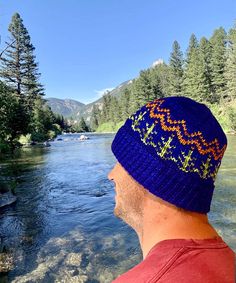 a man wearing a blue knitted beanie looks out over a river in the mountains