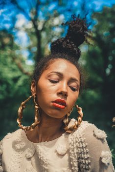 a woman with her eyes closed wearing large gold hoop earrings and a white dress in front of trees