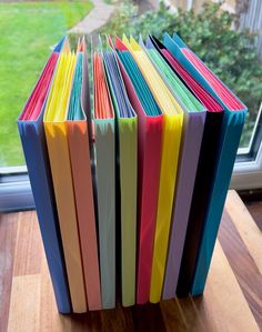 a stack of colorful books sitting on top of a wooden table next to a window