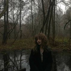 a woman standing in front of a body of water surrounded by trees and leaves on a foggy day