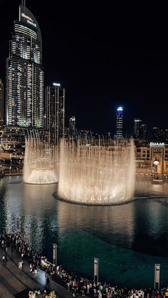 the water fountain is lit up at night in front of cityscape and skyscrapers