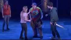 three people standing on stage talking to each other and one person holding a large knife