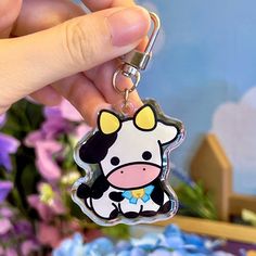 a hand holding a cow shaped keychain in front of purple and blue flowers