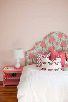 a bed with pink and green pillows on it next to a white nightstand table in front of a pink wall