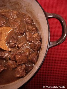 a pot filled with beef and sauce on top of a red table cloth next to a wooden spoon