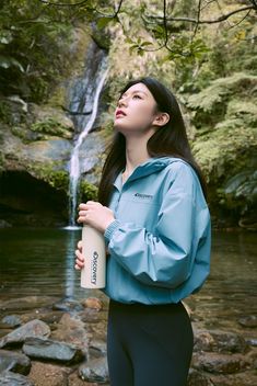 a woman standing in front of a waterfall holding a water bottle and looking up into the sky