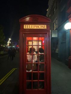 two people in a red phone booth on the street at night with one person taking a photo