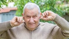 an older man is holding his hands to his ears and looking at the camera while standing outside