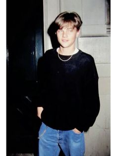 a young man standing in front of a door wearing jeans and a black sweater with a pearl necklace