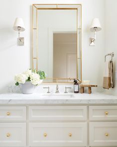 a white bathroom with marble counter tops and gold framed mirror above the sink, along with flowers