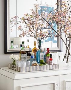 a white dresser topped with lots of bottles and glasses next to a vase filled with flowers