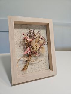 a frame with dried flowers in it sitting on a white table top next to a wall