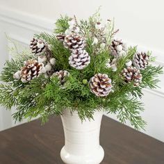 a white vase filled with pine cones and greenery on top of a wooden table