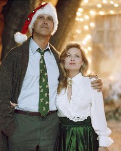 a man standing next to a woman wearing a santa hat and green skirt in front of christmas lights