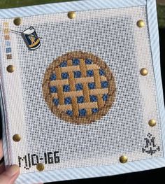 a hand holding up a cross stitch picture frame with a pie on the front and bottom