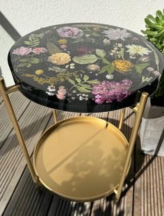 a black and gold side table with flowers painted on it, next to a potted plant