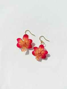 two red flowers are hanging from gold earwires on a white surface, one is pink and the other is orange