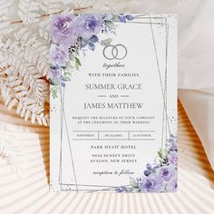 a wedding card with purple flowers and two rings on it next to some white feathers