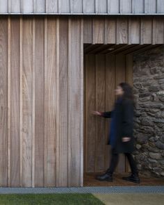 a woman walking out of a wooden door into a stone building next to a grass covered field