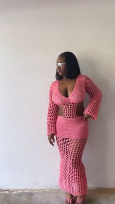 a woman in a pink crochet dress standing against a wall with her hands on her hips