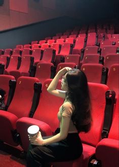 a woman sitting on a red chair in front of an empty theater seat holding a cup