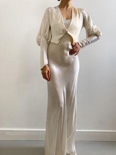 a woman wearing a white dress and jacket standing in front of a wall with her hands on her hips