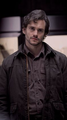 a man standing in front of a wall wearing a brown shirt and black jacket with his hands in his pockets