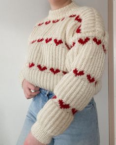 a woman wearing a white sweater with red hearts on the sleeves and arms, standing in front of a wall