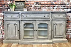 an old gray painted buffet with glass doors and shelves on the sideboard in front of a brick wall