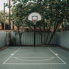 an empty basketball court in front of a fence with trees and bushes around it,