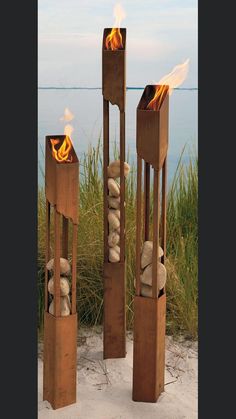 three wooden sculptures with fire in them on the sand next to some grass and water