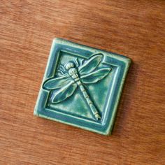 a ceramic tile with a dragonfly on it's side sitting on a wooden surface