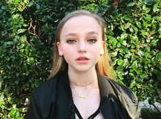 a young woman standing in front of a bush wearing a black jacket and gold necklace