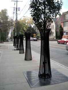 three tall black metal vases sitting on the side of a sidewalk next to a street