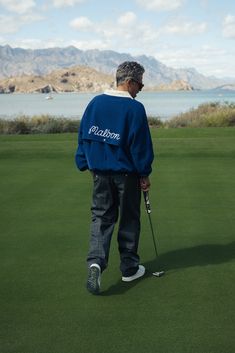 a man in a blue jacket is playing golf on the green with mountains in the background