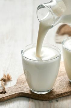 milk being poured into two glasses on top of a wooden board