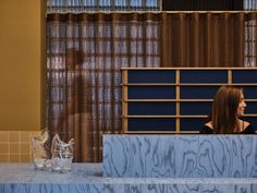 a woman sitting at a table in front of a glass vase and book shelf with books on it