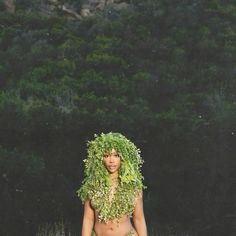 a woman with green hair and grass on her head is standing in front of trees