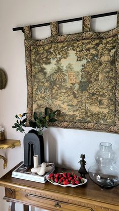 a table topped with a bowl of strawberries on top of a wooden table next to a tapestry