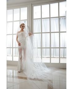 a woman in a white wedding dress standing by a window with her veil pulled back