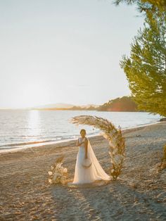 a woman in a wedding dress standing on the beach next to an arch made out of flowers