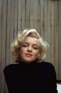 a woman with blonde hair sitting in front of a wooden wall wearing a black top