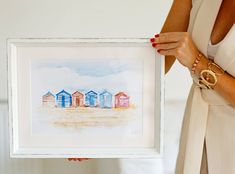 a woman holding up a watercolor painting of beach huts