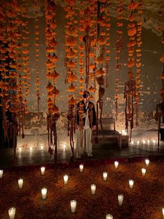 a bride and groom standing in front of many lit candles at their wedding ceremony with orange flowers hanging from the ceiling