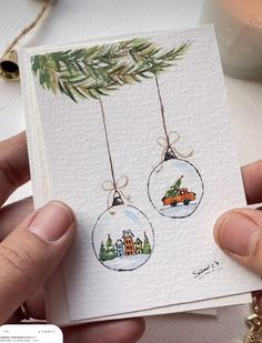 a hand holding a christmas card with two ornaments hanging from it's sides and a pine tree in the background