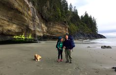 a man and woman standing on the beach with a dog in front of some cliffs