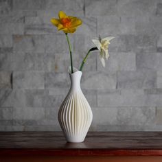 two yellow and white flowers are in a vase on a wooden table next to a brick wall