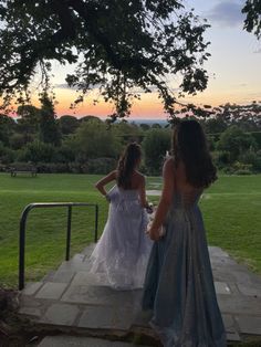 two women in dresses are standing on a stone platform under a tree and looking at the sunset